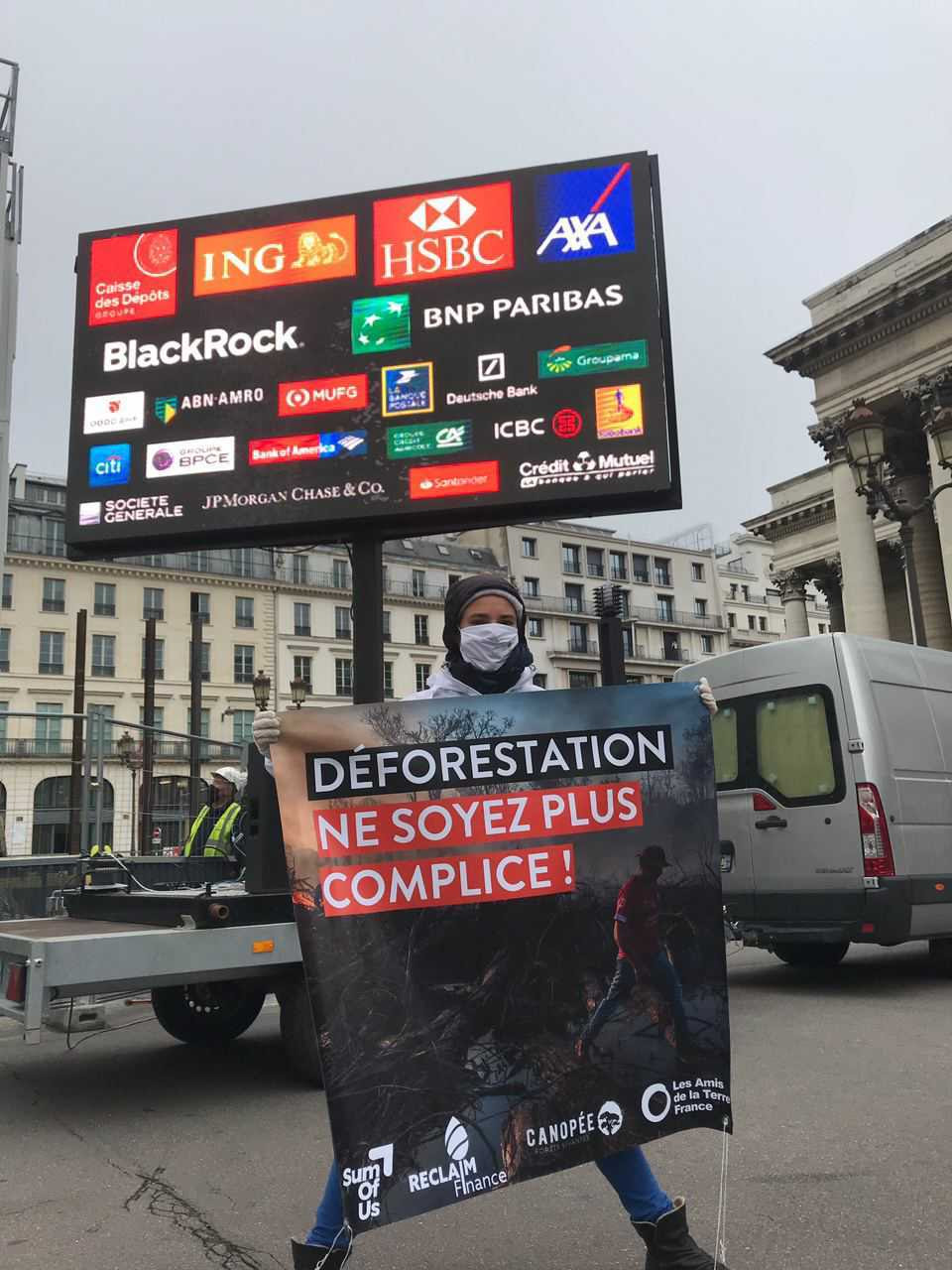 Activist holding a banner "Deforestation: Stop complicity", in front of the billboard broadcasting the names of the financial actors involved.
