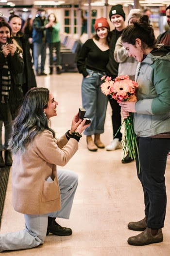 Person on one knee opening a small box with a ring in it as another stands up holding flowers and several people in the background watching