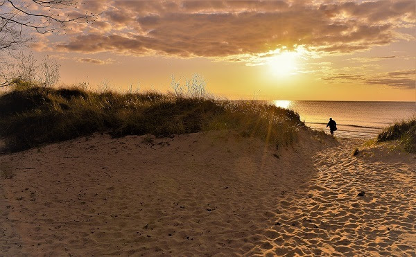 a man in shadow walks the sandy beach toward a huge, flat lake, as golden sun breaks through just above low-set clouds