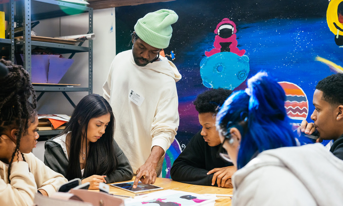 Brandon Breaux, a Black man with a deep skin tone, stands over five seated students. On the table is an iPad and illustrations. He is wearing a mint beanie. Space art is on the wall in the background. 