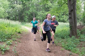 a small group of young girls and boys, clothed in shorts, T-shirts and sweatshirts, smile while running down a dirt path lined by mature trees