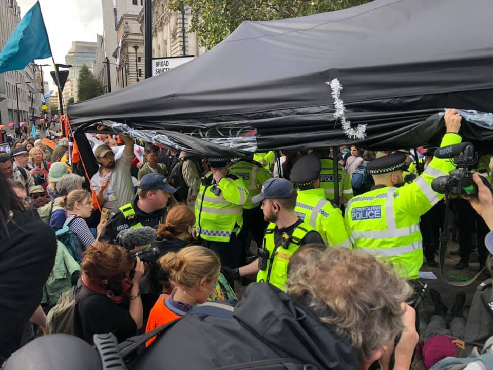 A group of around 10 police officers attempting to seize a black gazebo, with rebels and people with cameras surrounding them.