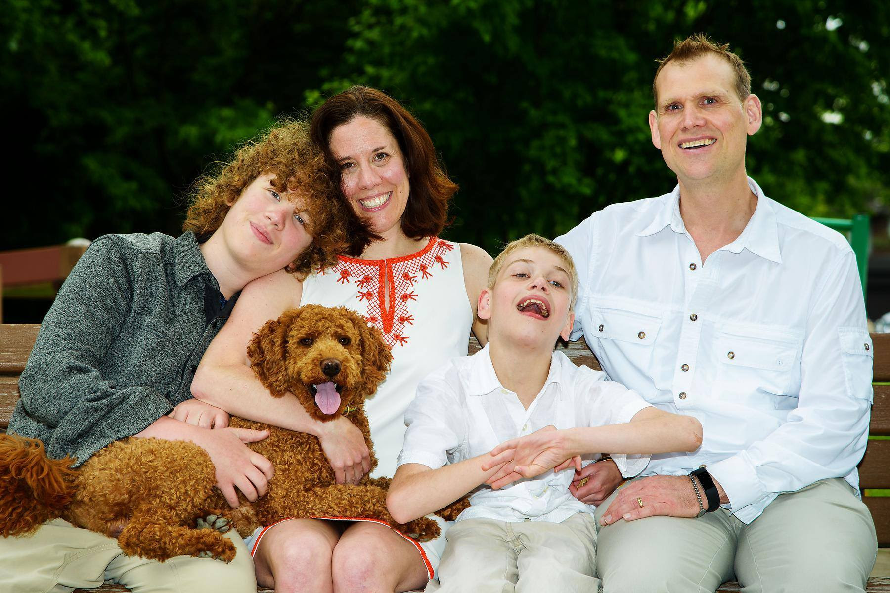 A family sitting on a bench. One son on the left is having curly red hair and a cute labradoodle on his lap. Next to him is his mom, a woman with brown hair who smiles brightly. Next to her,
the younger son who wears a white shirt. And on the right side is the dad wearing a white shirt as well. 