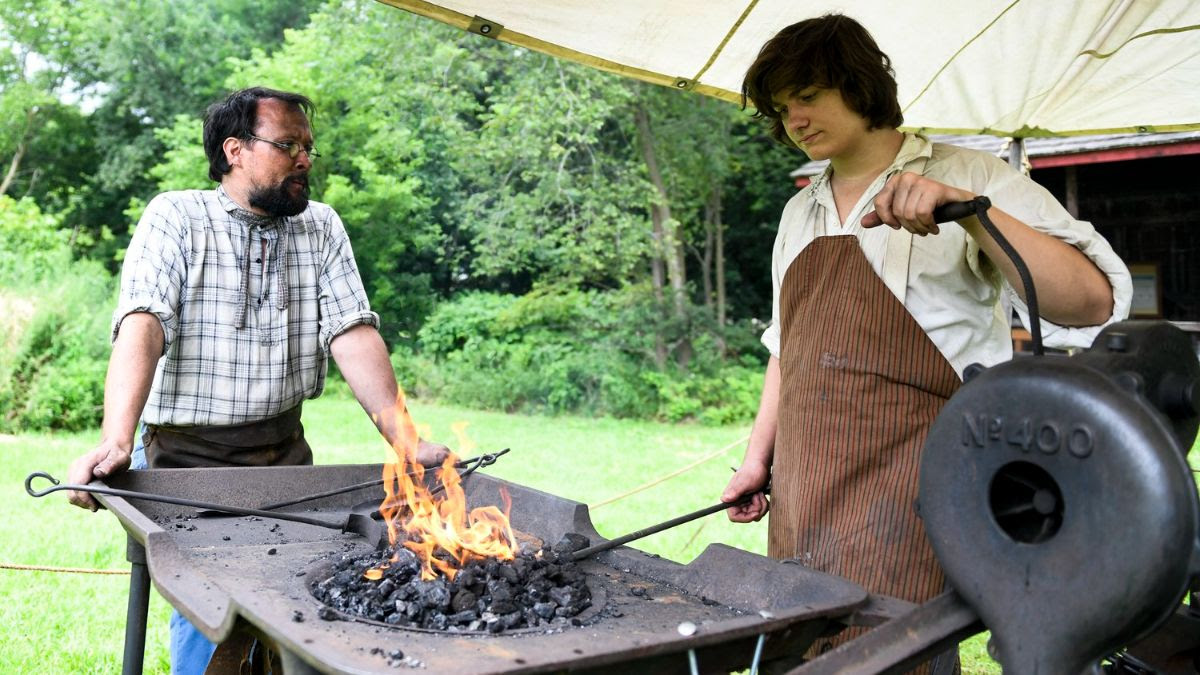 A man dressed in historical costumer guides a boy, also in historical costume, working at a blacksmith forge.