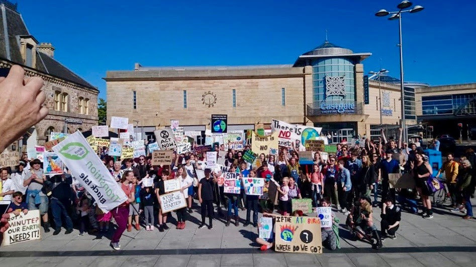 A group shot of 100+ strikers in a town square, holding signs with a building and blue sky behind them.