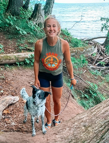 a smiling woman in workout clothing on a tree-lined trail, holding the leash of a medium-sized, fluffy black and white dog, lake in background