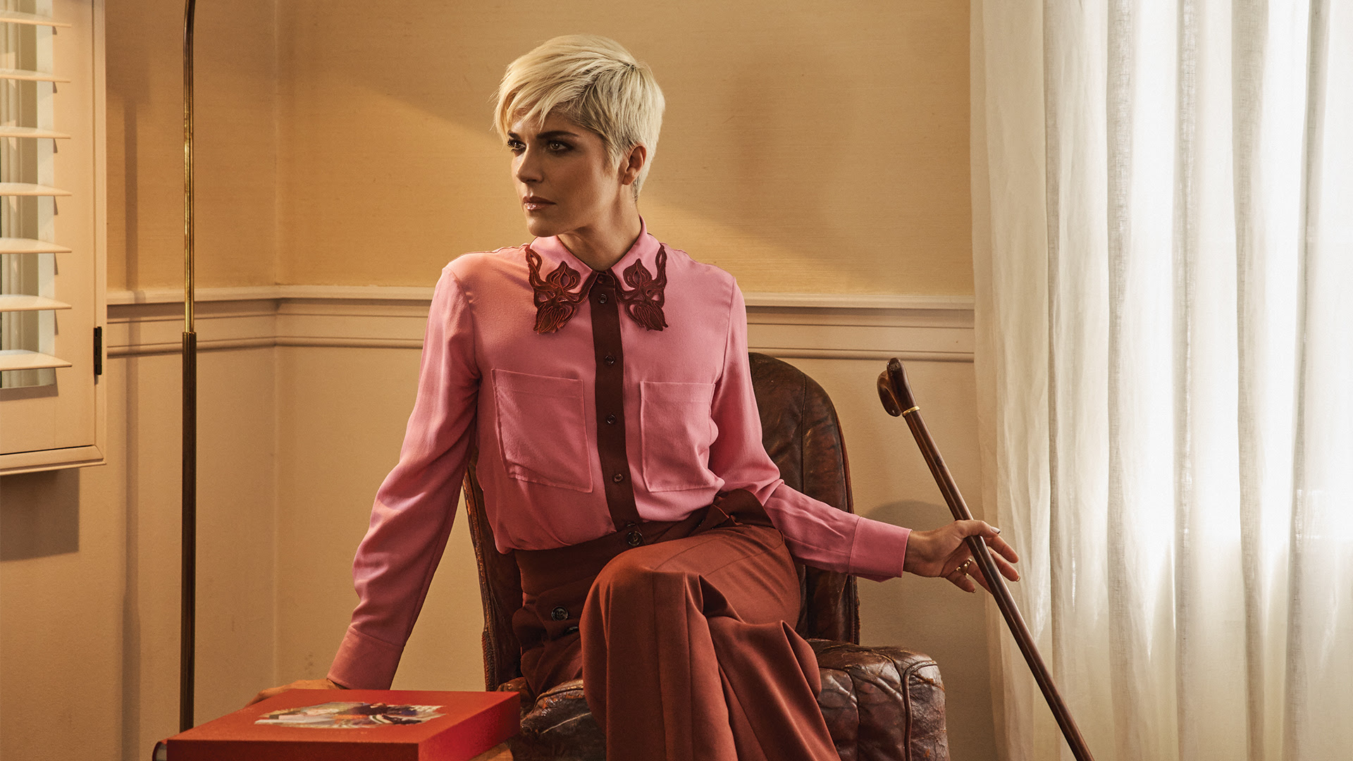Selma Blair, a woman with short blonde hair sits on a chair with her walking cane in one hand. She is dressed in purple and red