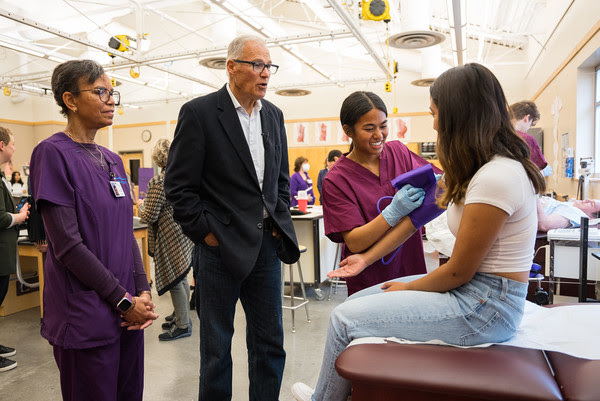 Gov. Jay Inslee watches students perform health care practice exercises in a classroom.