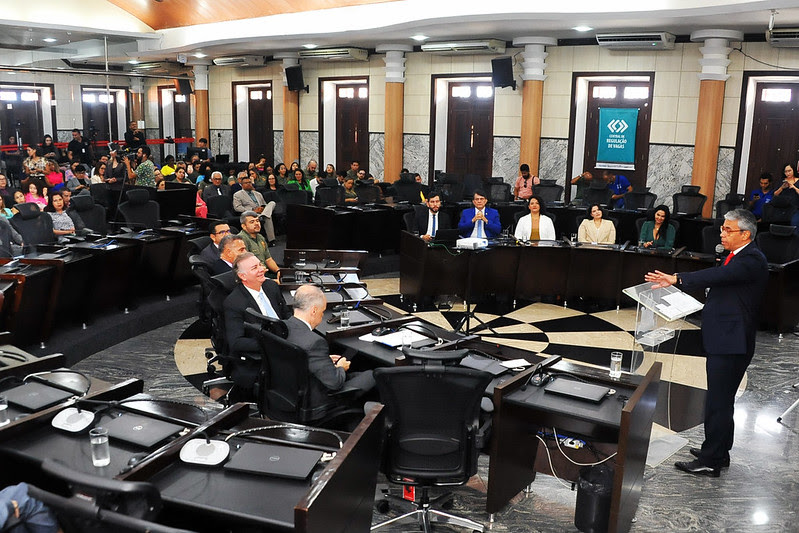 Foto no plenário do TJMA durante solenidade, com mesa de autoridades em formato circular e plateia ao fundo, e presidente do TJMA à frente, fazendo uso da palavra.