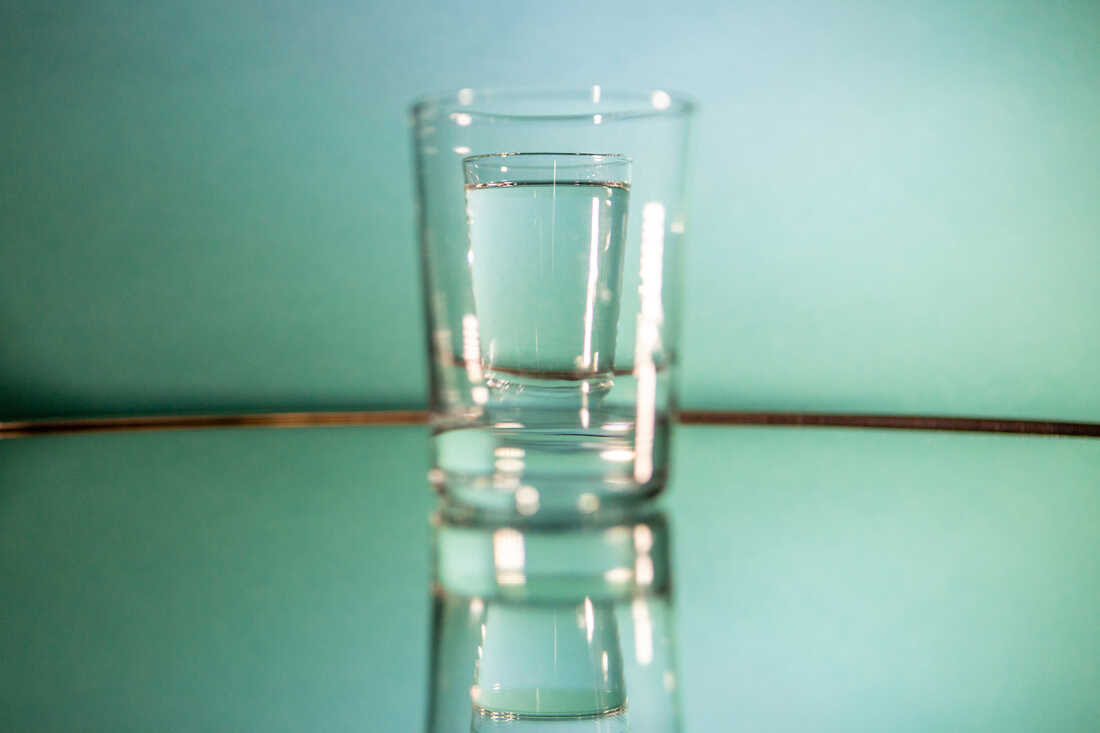 A full glass of water is photographed through an empty glass of water in the foreground so that the full glass of water appears smaller inside of the empty one. The two glasses sit on a mirror with a light green backdrop behind them.