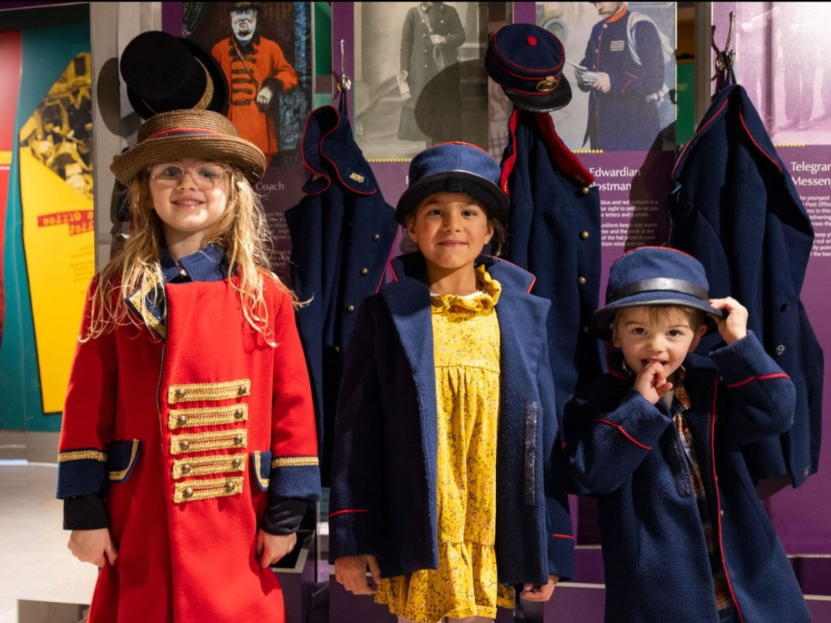 Marketing Photograph: Three children stand in the mueum galleries facing the camera playing dress up in different postal worker uniforms 