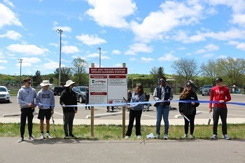 Seven people stand next to a sign on the edge of a parking lot preparing to cut a ribbon held in front of them.