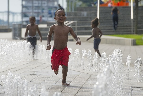 a smiling young boy in navy blue swim trunks jumps in the spray of a concrete splashpad, with two small boys playing behind him