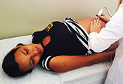 Pregnant teenage girl lies on her back in an exam room while a medical worker with her back to the camera examines her belly