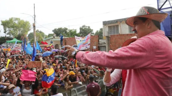 A man in a red button down shirt addresses a large crowd, signs and Venezuelan flags being held overhead.