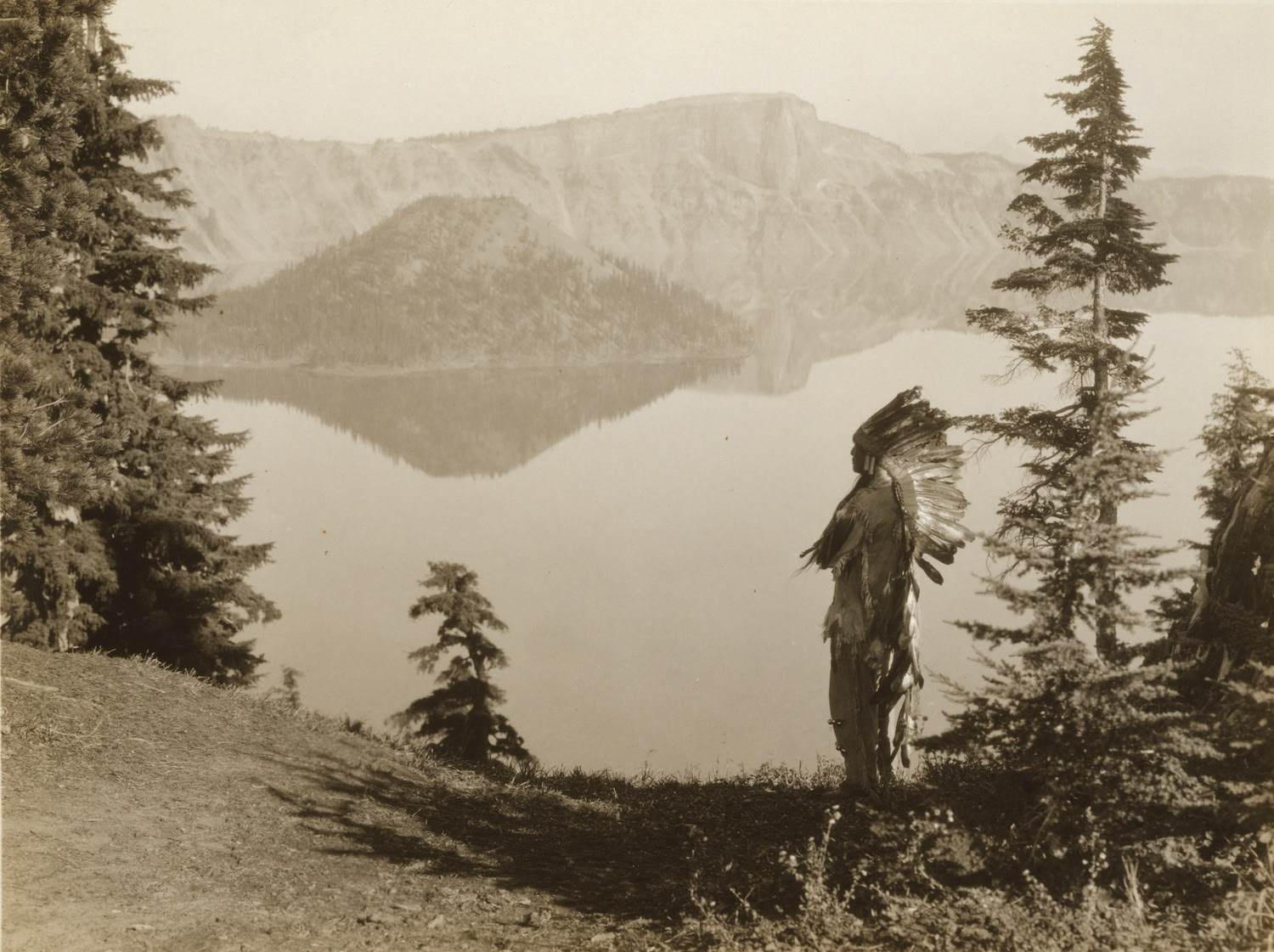Klamath Indian chief in ceremonial headdress standing on hill overlooking lake, California or Oregon. November 2021