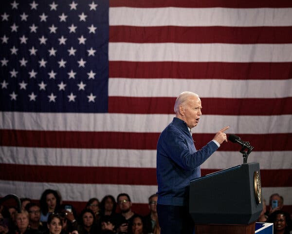 President Biden standing at a podium speaking at a campaign event with a large American flag hanging in the background.