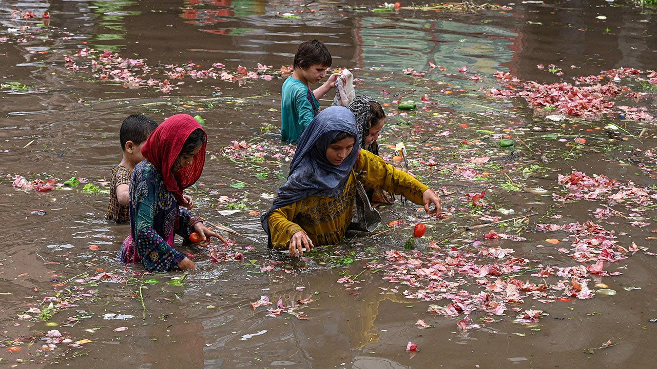 Five children, three wearing headscarves, are seen in waist-deep floodwaters with floating debris.