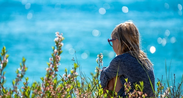 dark-blond-haired little girl wearing glasses, knees pulled up, sitting among tall green and pink stalked flowers, bright blue water in background