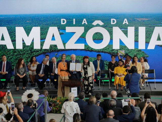 Cerimônia comemorativa do Dia da Amazônia no Palácio do Planalto. Na imagem é possível ver um telão com a frase "Dia da Amazônia" sobreposta a uma imagem aérea da floresta. À frente estão ministros do governo Lula sentados em cadeiras, com Lula, Joenia Wapichana e Marina Silva em pé diante do público ouvinte segurando um documento