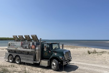 A dark green and silver DNR fish stocking truck, with the words Fish for the Future on the side, on sandy shore near wide river mouth