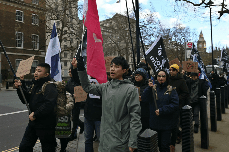 A group of political activists bundled up in warm clothing walks along a sidewalk in London while protesting a law in Hong Kong. Some of the demonstrators hold flags and handwritten cardboard signs.