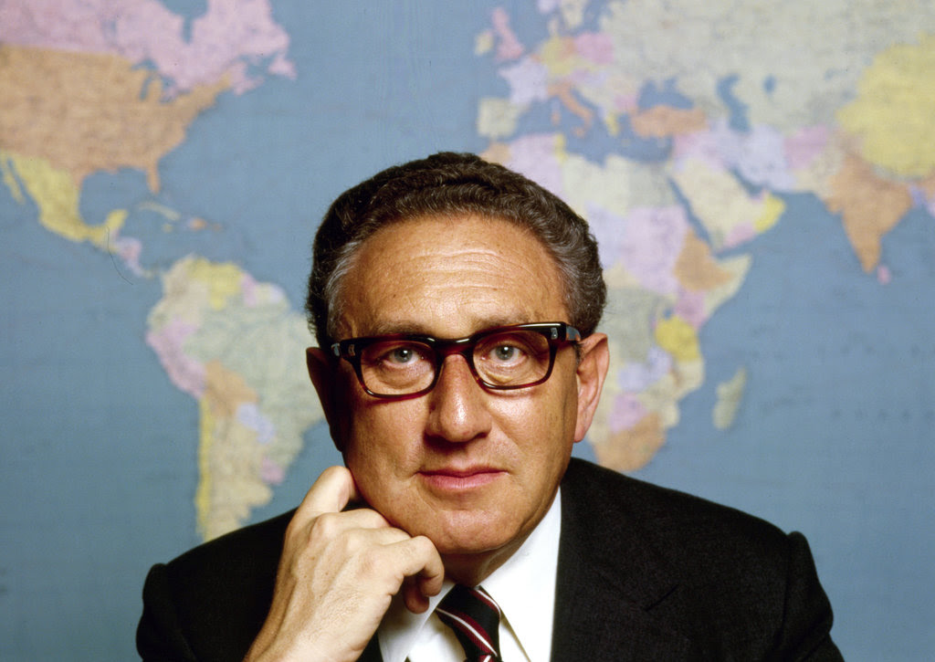 A color portrait of Ms. Kissinger in a dark suit jacket, white shirt and tie, his right hand pressed against his chin. A wall map of the world fills the space behind him.