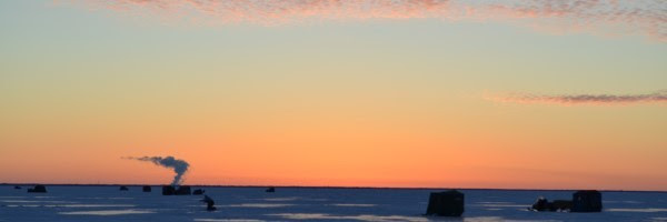 A soft orange sunset spreads over an iced-over lake with the dim outlines of ice fishing shanties dotting the skyline.