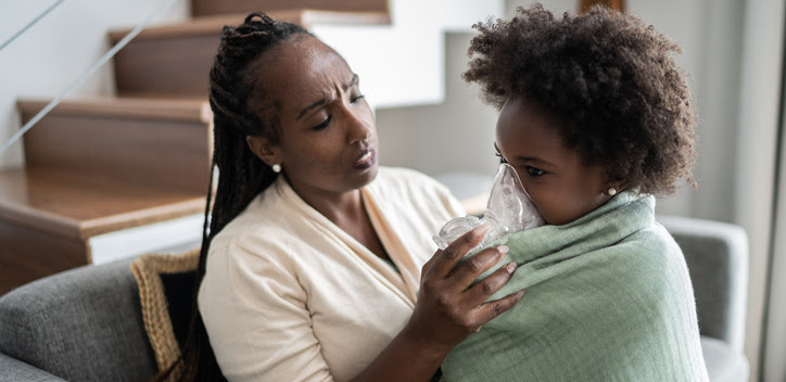 A worried Black mother sits in her living room, holding an oxygen mask to her young daughter's face.