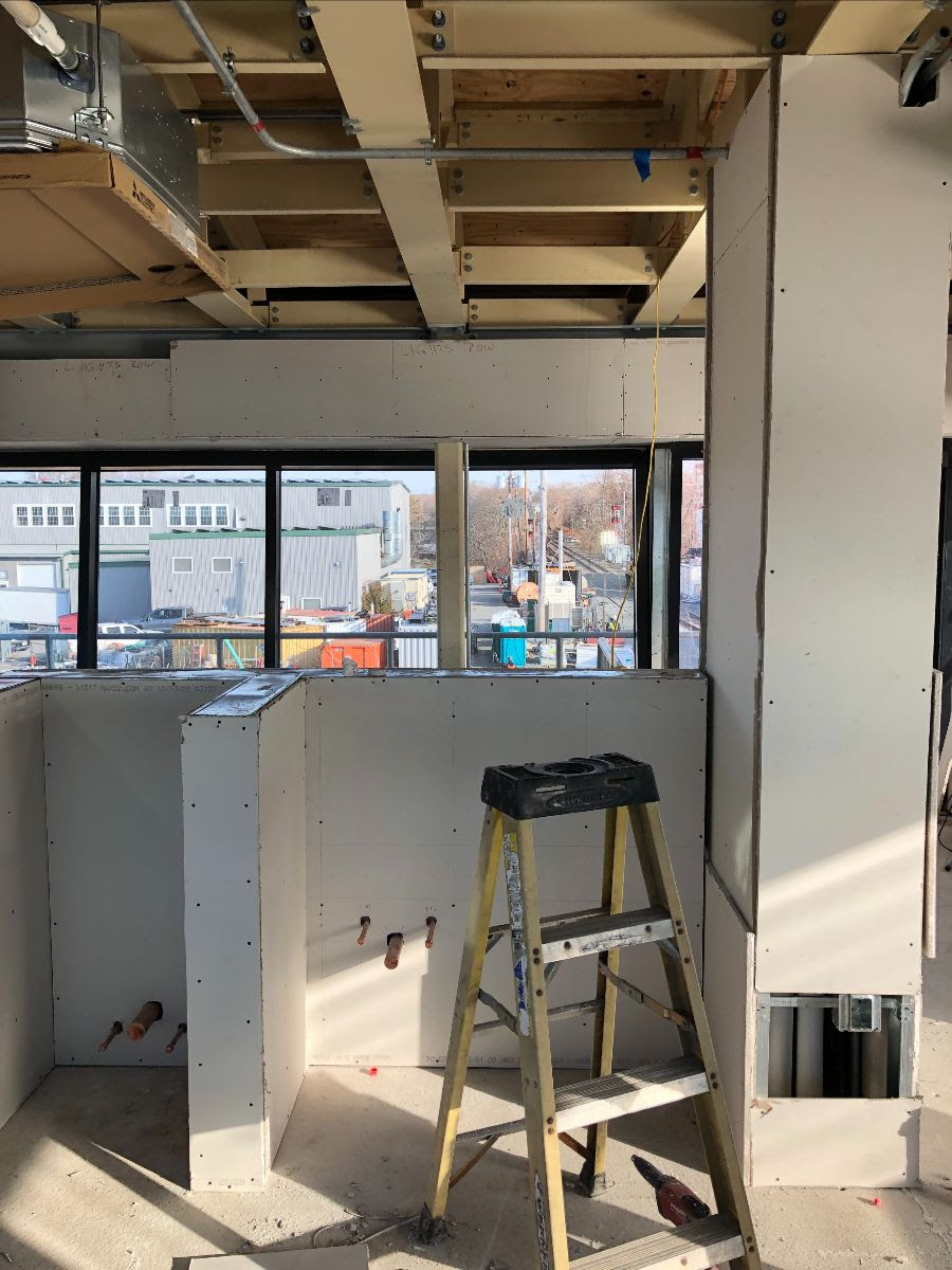 A view of the interior of the control tower facing toward the windows shows that drywall has been installed on walls and to create bays within the room. The ceiling has no drywall and is open so that rafters and beams are visible. 