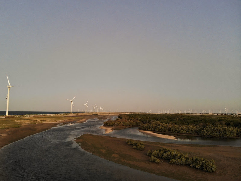 Ao por do sol, imagem de aerogeradores (torres brancas com hélices brancas na parte de cima), produzindo energia eólica em paisagem em faixa de areia no litoral, ao lado do estuário de rio.