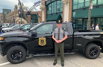 A uniformed, male conservation officer stands in front of his black DNR patrol truck, next to Comerica Park baseball stadium in Detroit