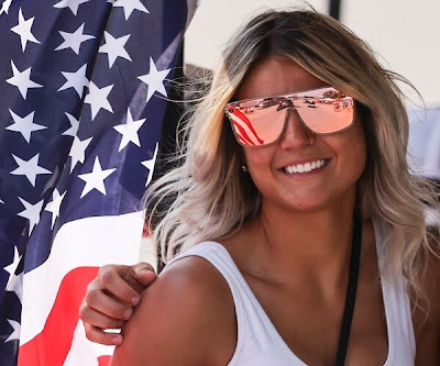 woman wearing mirror sunglasses stands next to american flag