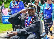 A Veteran wearing a sea captain's hat salutes while rolling his motorized wheelchair among others walking around him A Veteran wearing a sea captain's hat salutes while rolling his motorized wheelchair among others walking around him