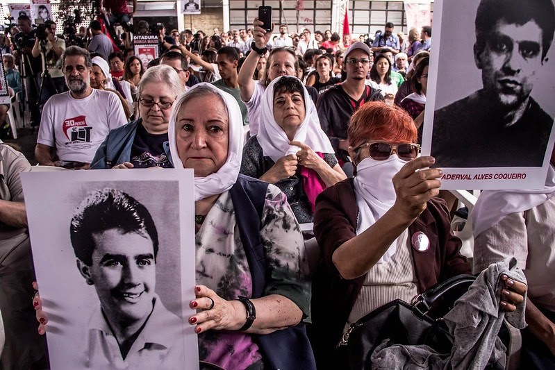 Familiares de mortos e desaparecidos na ditadura seguram cartazes com as fotos de seus parentes e o nome deles em vermelho