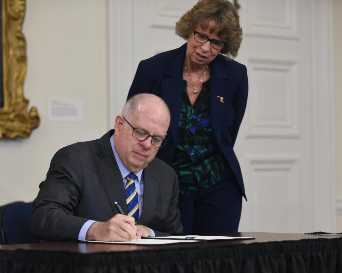 Secretary Beatty stands behind Governor who is seated as he signs executive order. 