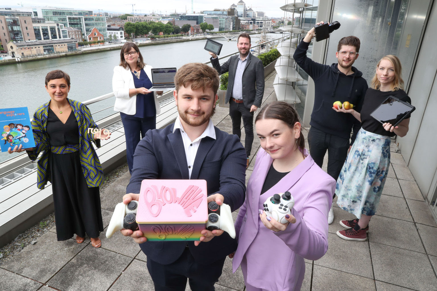 Two people standing outdoors with many people in the background. To the right is a woman in a pink jacket holding a gaming console and on the left is a man with curly brown hair holding a pink box saying ByoWave