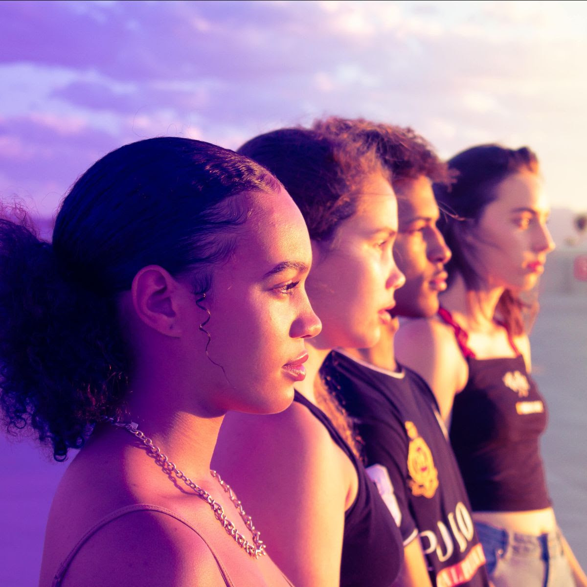 Side profile photo of four diverse young adults shoulder to shoulder outdoors with a cloudy background.