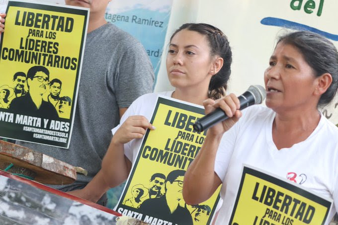 Vidalina Morales, environmental defender and president of ADES, speaking into a microphone while holding a sign calling for the release of the five detained environmental defenders. She is beside people who are holding a similar sign.