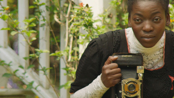 woman stands by green branches behind old camera