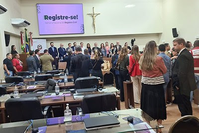 Em um salão com inúmeras pessoas em pé, se vê, em primeiro plano, algumas mesas de vidro com cadeiras. Na parede do fundo, se vê um telão com a mensagem "registre-se" e, ao lado dele, um crucifixo.