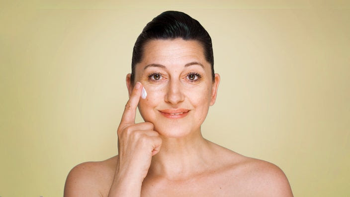 Studio portrait of a woman with short hair putting on anti-aging cream. The background is a gradient yellow color.