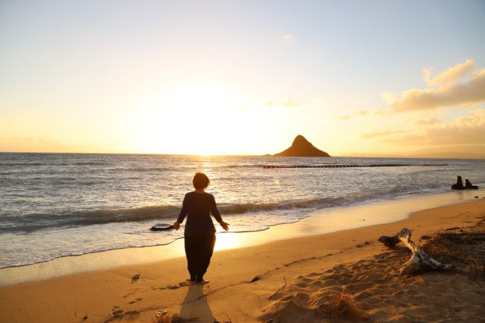 siloette image of Emma Kupu Mitchell with the sun rising behind her on a beach with an island mountain in the background