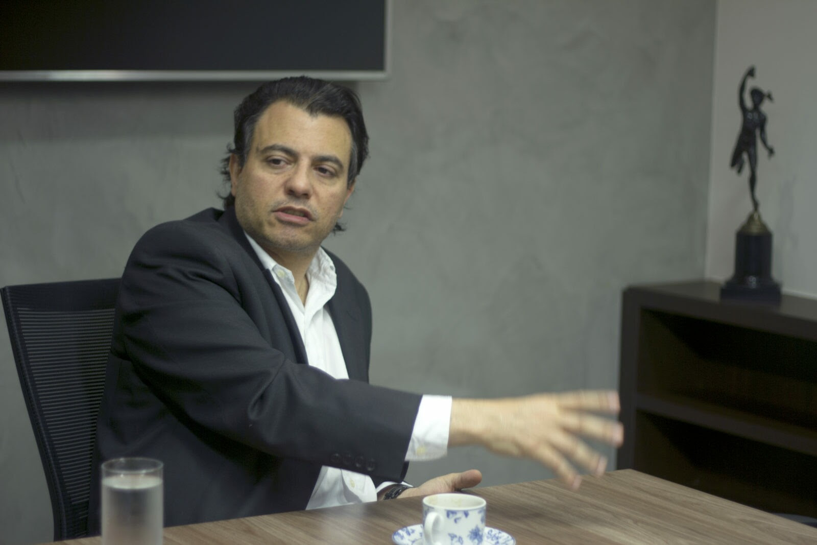 Otávio Fakhoury é um homem branco com cabelos escuros; na foto ele veste um blazer preto com uma camisa branca; ele está no seu escritório em São Paulo