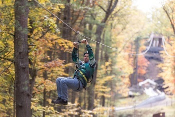 a man in safety gear and helmet and strapped to a zip line, holds on to the handles as he rides through a colorful autumn forest