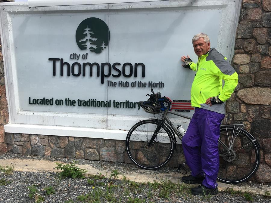 Man stands besides bicycle in front of the city sign of Thompson.