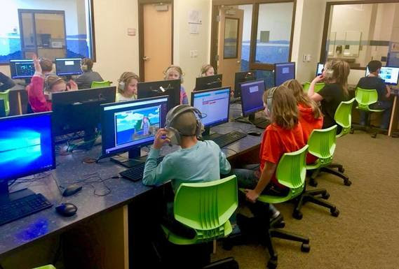 A classroom full of elementary students sit at computers playing games that teach coding.