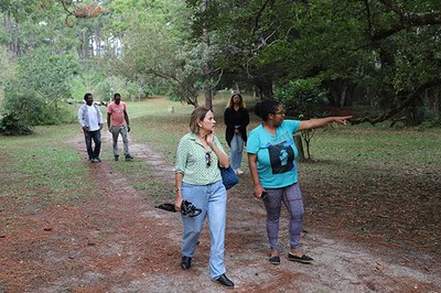 Cinco pessoas caminham em uma trilha, cercada de vegetação. Duas mais à frente e as outras três atrás delas. Uma das pessoas mais à frente aponta o dedo para algo que está fora do enquadramento da foto.