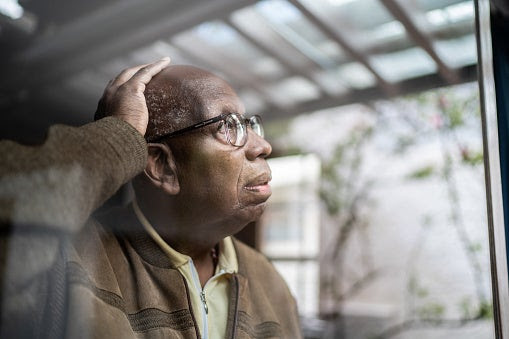 Portrait of an elderly man looking out of a window with his hand on his head.