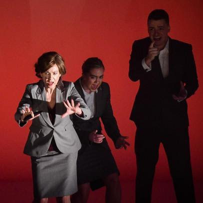 Three figures in suits on a stage with a red backdrop